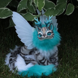 Op de afbeelding: Een witte en grijze harige kat met blauwe vleugels en geweien. De kat heeft blauwe ogen en een turquoise blauwe kraag. De kat zit op groen gras.