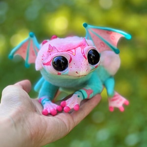 Può includere: Un piccolo drago di peluche rosa e blu con grandi occhi neri e ali. Il drago è seduto su una mano.