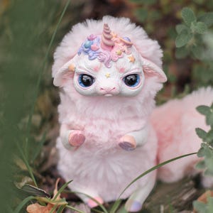 May include: A pink fluffy unicorn doll with a colorful horn and a galaxy-themed design on its head. The doll has large blue eyes and is holding two pink and white objects in its paws.