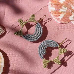 Può includere: Un paio di orecchini a cerchio argentati e dorati con un design intrecciato unico. Gli orecchini sono esposti su una superficie rosa con conchiglie.