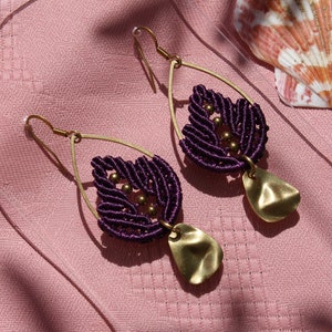 May include: A pair of handmade earrings featuring a dark purple macrame design with gold-colored accents and teardrop-shaped pendants. The earrings hang from gold-colored ear wires. The earrings are displayed on a pink surface with seashells.