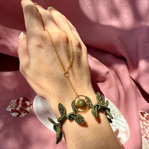 May include: A handmade macrame bracelet in olive green with a gold chain and a gold ring holding a green bead. The bracelet is worn on a wrist. The background is a pink fabric.