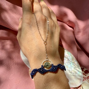 May include: A handmade blue macrame bracelet with a silver chain and a green and gray stone. The bracelet is connected to a silver chain that goes up to a ring. The jewelry is displayed on a pink background.