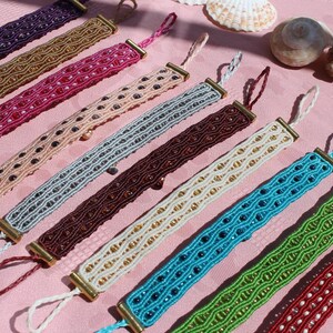 May include: A collection of colorful macrame bracelets in various shades, including purple, gold, pink, and blue, with decorative beads and gold-toned metal accents. The bracelets are displayed on a pink surface, with seashells in the background.
