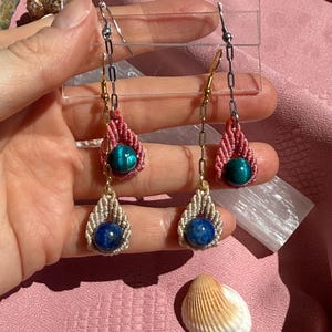 May include: Dangle earrings featuring a silver-toned chain connected to a macrame-knotted teardrop shape. Each earring holds two beads: a teal stone bead in a pink macrame setting and a dark blue stone bead in a beige macrame setting.