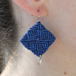 May include: Blue and silver square-shaped earrings with a crystal pendant. The earrings are made with a knotted cord and have a geometric design.