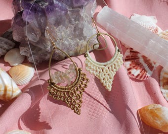 May include: Two pairs of handmade earrings with intricate macrame detailing. One pair is a light beige color, the other is a golden brown. The earrings are displayed on a pink fabric with seashells, crystals, and a selenite wand.