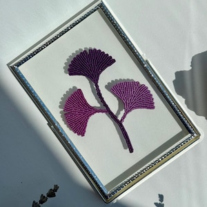 May include: A framed artwork featuring a purple ginkgo leaf design. The leaves are intricately crafted with a textured appearance, set against a white background within a silver-edged frame. The artwork is displayed on a white surface.