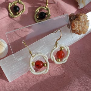 May include: Two pairs of dangle earrings featuring circular woven off-white cord frames. Each earring contains a central round bead, with red beads in the foreground and black beads in the background, accented by small metallic gold-toned spheres.