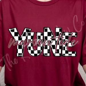May include: Maroon-colored t-shirt with the text "YUNE Maroon Tide" in a black and white checkered pattern. The shirt has a crew neck and short sleeves. The text is in a bold, blocky font with a cursive font for "Maroon Tide".