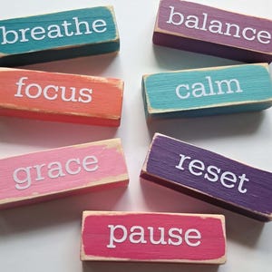 May include: A collection of rectangular wooden blocks painted in various colors, including teal, orange, pink, purple, and turquoise. Each block features a white word: "breathe," "focus," "grace," "balance," "calm," "reset," and "pause."