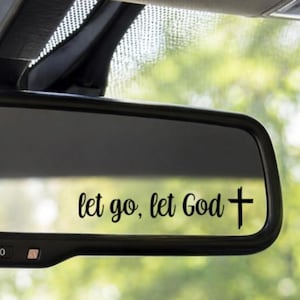 May include: A car's rearview mirror with the text "let go, let God" in a script font, followed by a cross symbol. The mirror is black, with a blurred green background visible through the windshield.