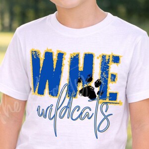 以下が含まれることがあります： 青と金色の「WHE Wildcats」の文字と足跡のグラフィックが入った白いTシャツ。屋外で笑顔の子供が着ています。