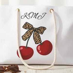 Puede incluir: Bolso tote blanco con asas de cuerda y diseño de cerezas. El bolso presenta un lazo con estampado de leopardo y dos cerezas rojas. Las iniciales "KMJ" están impresas encima del diseño. Un accesorio elegante y práctico.