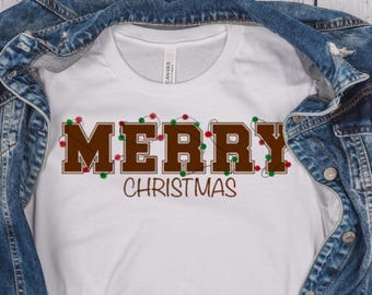 Op de afbeelding: Wit T-shirt met de tekst "MERRY CHRISTMAS" in bruine blokletters, versierd met rode en groene kerstverlichting. Het T-shirt wordt getoond op een spijkerjasje.