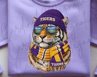 May include: A purple sweatshirt featuring a tiger wearing a purple beanie with "TIGERS" in white, aviator sunglasses, and a purple and gold scarf with "TIGERS". The tiger's face is the focal point, set against a plain purple background.