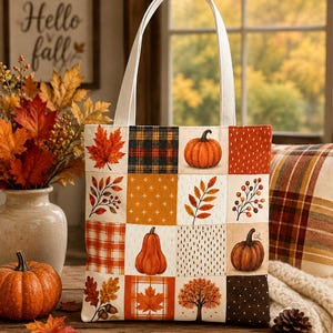 May include: A tote bag featuring a patchwork design of various autumn-themed squares, including illustrations of pumpkins, maple leaves, acorns, a tree, and plaid patterns. The bag has two white fabric handles and a rectangular, flat-bottomed shape.