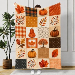 May include: A rectangular throw blanket featuring a patchwork grid design with autumn-themed illustrations. The pattern includes orange pumpkins, red and yellow maple leaves, brown acorns, bare trees, and various plaid, dotted, and cross-stitched textures.