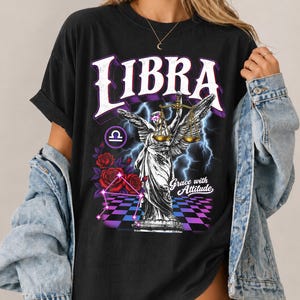 Könnte beinhalten: Ein schwarzes T-Shirt mit einem Grafikdruck, der das Wort LIBRA in großen weißen Buchstaben zeigt. Das Design umfasst eine geflügelte Statue mit Waage, rote Rosen, blaue Blitze, einen violett-schwarzen Schachbrettboden und den Text Grace with Attitude.