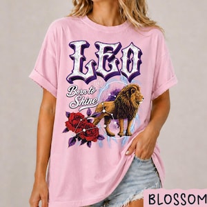 Op de afbeelding: Een lichtroze T-shirt met korte mouwen en de tekst LEO en Born to Shine in een donkerpaars lettertype. De afbeelding bevat een bruine leeuw, twee rode rozen met groene bladeren en een wit sterrenbeeld op een paarse cirkelvormige achtergrond.
