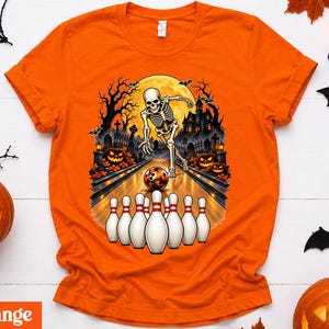 Könnte beinhalten: Ein orangefarbenes T-Shirt mit einem Grafikdruck eines Skeletts, das auf einer Bahn in einem Friedhofsbereich bowlt. Die Szene zeigt einen Vollmond, ein Spukhaus, Kürbisse, Fledermäuse und eine Reihe weißer Bowling-Pins mit roten Streifen.
