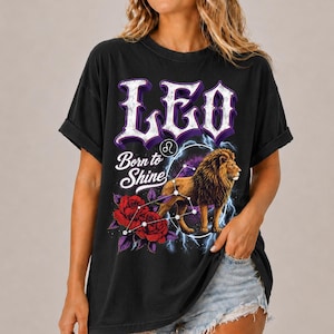 Op de afbeelding: Een zwart t-shirt met korte mouwen en een grafische print met de tekst LEO en Born to Shine. Het ontwerp bevat een leeuw, rode rozen, een sterrenbeeld-symbool en blauwe bliksemschichten tegen een donkere achtergrond op de voorzijde.