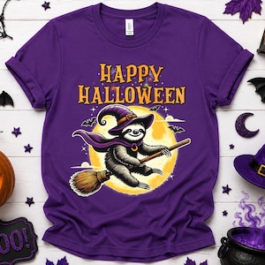 May include: A purple short-sleeved t-shirt featuring a graphic of a sloth wearing a purple witch hat and cape, flying on a broomstick in front of a large yellow moon. The text "HAPPY HALLOWEEN" is printed in orange above the sloth illustration.
