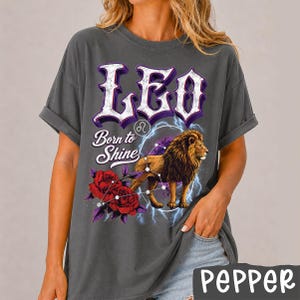Op de afbeelding: Een antracietgrijs T-shirt met korte mouwen en een grafische print met de tekst LEO en Born to Shine. Het ontwerp bevat een leeuw, rode rozen, een sterrenbeeld-symbool en blauwe bliksemschichten. Het shirt heeft een relaxte pasvorm en opgerolde mouwen.