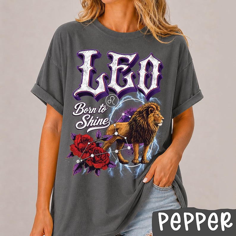 Op de afbeelding: Een antracietgrijs T-shirt met korte mouwen en een grafische print met de tekst LEO en Born to Shine. Het ontwerp bevat een leeuw, rode rozen, een sterrenbeeld-symbool en blauwe bliksemschichten. Het shirt heeft een relaxte pasvorm en opgerolde mouwen.