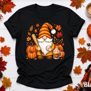 Könnte beinhalten: Ein schwarzes Kurzarm-T-Shirt mit einer Grafik eines Gnoms mit gestreifter Mütze, der einen Baseballschläger und einen Baseball hält. Der Gnom ist umgeben von orangefarbenen Kürbissen, Herbstblättern und einer Tasse Kaffee mit Sahne.