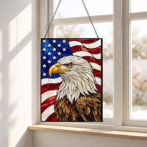 Könnte beinhalten: Buntglasfenster mit einem Weißkopfseeadler und der amerikanischen Flagge. Der Adler hat weiße Federn, einen gelben Schnabel und braune Augen. Die Flagge hat rote und weiße Streifen und ein blaues Feld mit weißen Sternen. Das Panel ist schwarz gerahmt und hängt an einer Kette.