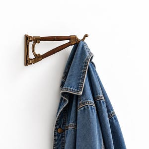 May include: A blue denim jacket hangs from a wall-mounted coat rack. The rack features a dark brown wooden arm with brass-toned metal hardware, including a curved support bracket and a hook at the end holding the jacket by its collar.