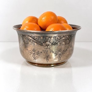 May include: A silver-toned metal bowl with a wide, flared rim and a rounded base, featuring intricate embossed floral and scrollwork patterns around the exterior. The bowl is filled with several bright orange, round citrus fruits.