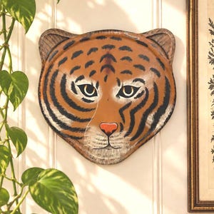 Könnte beinhalten: Ein dekoratives Wandobjekt in Form eines Tigerkopfes mit einem orangefarbenen Gesicht, schwarzen Streifen, weißen Akzenten um Augen und Schnauze sowie einer kleinen roten Nase. Der Tigerkopf hat abgerundete Ohren und eine glatte, gewölbte Oberfläche.