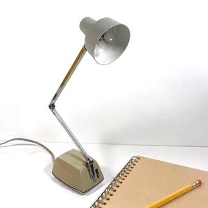 Puede incluir: Una lámpara de escritorio beige y cromada con brazo ajustable y pantalla cónica, situada junto a un cuaderno de espiral con cubiertas de papel marrón y un lápiz de madera amarillo con goma rosa sobre el cuaderno.