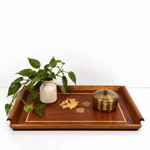 May include: A rectangular wooden serving tray featuring a central floral inlay design, accompanied by a small cream-colored ceramic vase with green foliage and a round, lidded brass container with a metallic finish.