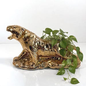 May include: A metallic gold-coloured ceramic planter shaped like a crouching tiger with its mouth open. The tiger has a textured, reflective surface and contains a green trailing plant with heart-shaped leaves spilling over the side of its back.