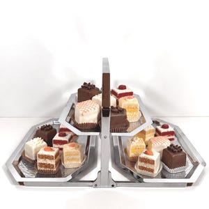 Può includere: Un'alzata a due ripiani in metallo argentato con manico centrale, contenente un assortimento di piccoli dolci quadrati. I dolci presentano varie guarnizioni, tra cui riccioli di cioccolato, frutti rossi e motivi di glassa bianca su fondo bianco.