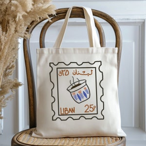 May include: A white canvas tote bag with a brown and black stamp design. The stamp features a blue and white coffee cup with steam coming from it. The text on the stamp reads "Liban 25p" in Arabic and English.
