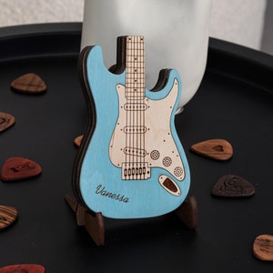 May include: A light blue miniature electric guitar with a wooden stand. The guitar has a white pickguard and is engraved with the name "Vanessa".