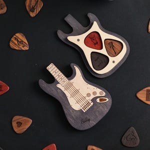 May include: Two wooden guitar-shaped boxes. One box is open and has three slots for guitar picks. The other box is closed and has the name "Billy" engraved on it. Both boxes are made of dark wood with a light wood interior.