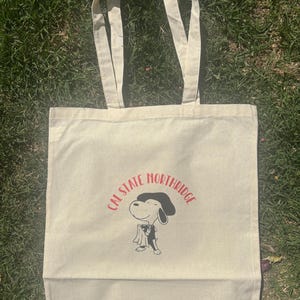Könnte beinhalten: Eine beige Canvas-Tragetasche mit rotem und schwarzem Text, der "Cal State Northridge" lautet. Ein Cartoon-Hund mit Hut und Smoking ist auf die Tasche gedruckt.
