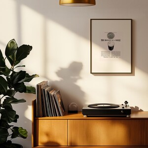 Puede incluir: Un tocadiscos negro con un disco de vinilo sobre un mueble de madera. Un grabado enmarcado con el texto "THE BEGINNING OF A SMILE" cuelga de la pared. Tambi&eacute;n se ve una pila de discos de vinilo y una planta en maceta.