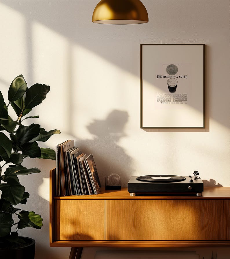 Puede incluir: Un tocadiscos negro con un disco de vinilo sobre un mueble de madera. Un grabado enmarcado con el texto "THE BEGINNING OF A SMILE" cuelga de la pared. Tambi&eacute;n se ve una pila de discos de vinilo y una planta en maceta.