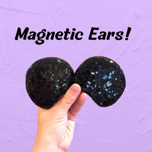May include: Two circular, black sequin-covered ears with a magnetic attachment feature. The text "Magnetic Ears!" is printed in black above the ears. The sequins have a reflective quality, showing subtle hints of blue and green light.