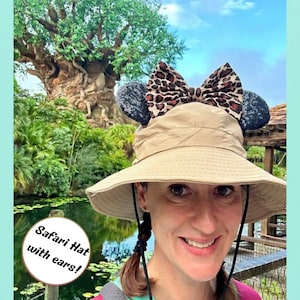 May include: A beige bucket hat featuring two black sequined ears and a large leopard print bow on top. The hat has a wide brim and a black chin strap. The text "Safari Hat with ears!" is printed in a circular graphic on the lower left side.