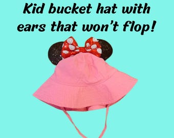 May include: A pink bucket hat for children featuring black circular ears and a red bow with white polka dots. The hat includes a chin strap and the text Kid bucket hat with ears that won't flop! printed in black above the hat on a light blue background.