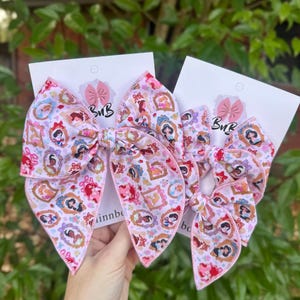 May include: Two pink hair bows with a princess-themed pattern. The bows are on white cards with a pink bow logo and the letters "BuB". The bows feature a repeating pattern of princess portraits in ornate frames.