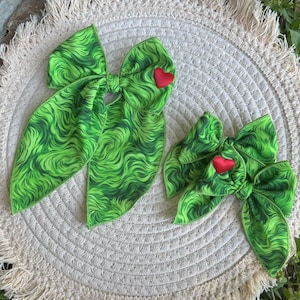 May include: Two green and red hair bows with a wavy pattern. The larger bow has a red heart in the centre and the smaller bow has a red heart on the right side.