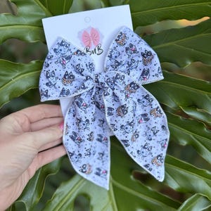 May include: White fabric hair bow with a repeating pattern of cartoon dogs in various poses. The bow is tied in the center and has a white border. The bow is on a white card with the logo "BuB".